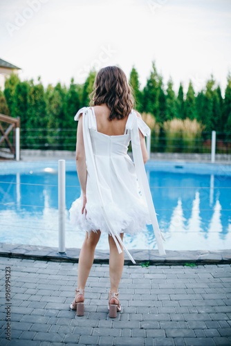 Young woman in white dress with feather hem posing near swimming pool, stylish summer portrait. Elegant feminine look, natural light, modern outdoor lifestyle scene with water reflections.