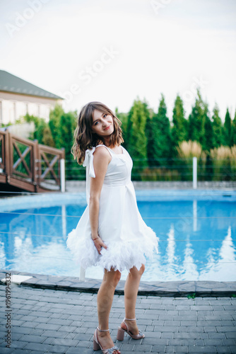 Young woman in white dress with feather hem posing near swimming pool, stylish summer portrait. Elegant feminine look, natural light, modern outdoor lifestyle scene with water reflections.