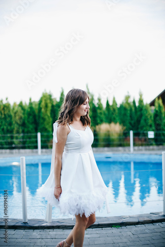 Young woman in white dress with feather hem posing near swimming pool, stylish summer portrait. Elegant feminine look, natural light, modern outdoor lifestyle scene with water reflections.