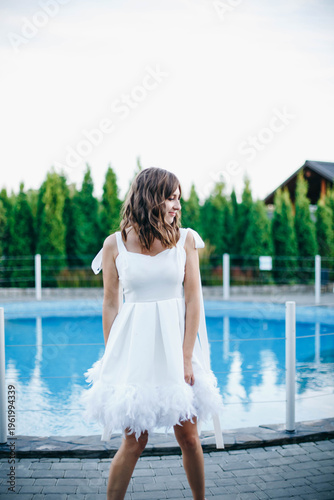 Young woman in white dress with feather hem posing near swimming pool, stylish summer portrait. Elegant feminine look, natural light, modern outdoor lifestyle scene with water reflections.