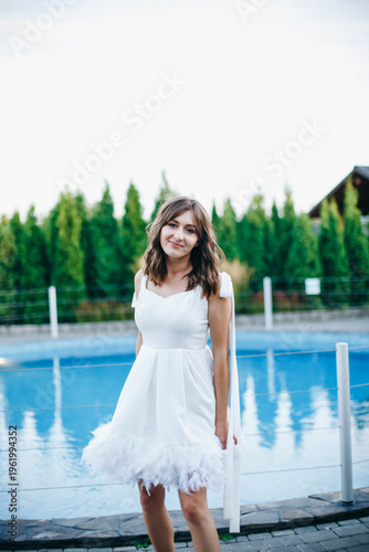 Young woman in white dress with feather hem posing near swimming pool, stylish summer portrait. Elegant feminine look, natural light, modern outdoor lifestyle scene with water reflections.