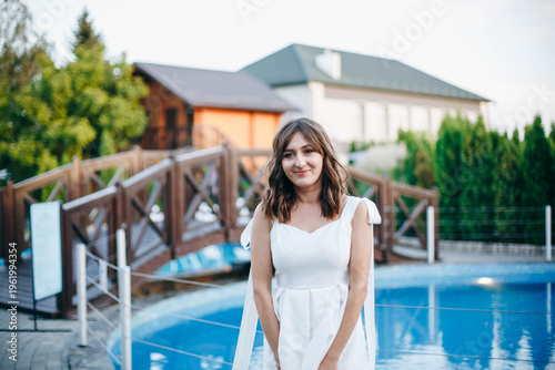 Young woman in white dress with feather hem posing near swimming pool, stylish summer portrait. Elegant feminine look, natural light, modern outdoor lifestyle scene with water reflections.