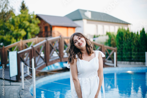 Young woman in white dress with feather hem posing near swimming pool, stylish summer portrait. Elegant feminine look, natural light, modern outdoor lifestyle scene with water reflections.