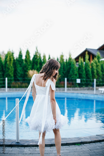 Young woman in white dress with feather hem posing near swimming pool, stylish summer portrait. Elegant feminine look, natural light, modern outdoor lifestyle scene with water reflections.