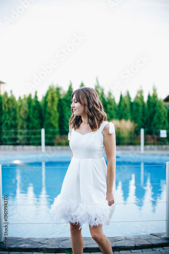 Young woman in white dress with feather hem posing near swimming pool, stylish summer portrait. Elegant feminine look, natural light, modern outdoor lifestyle scene with water reflections.