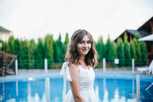 Young woman in white dress with feather hem posing near swimming pool, stylish summer portrait. Elegant feminine look, natural light, modern outdoor lifestyle scene with water reflections.