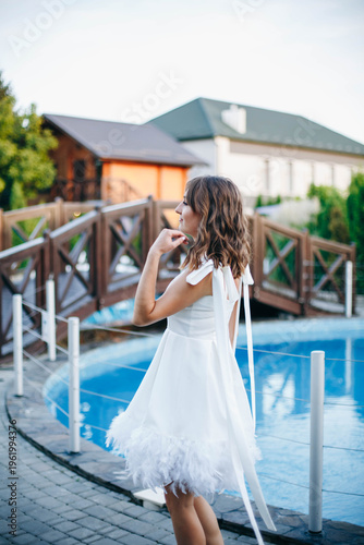 Young woman in white dress with feather hem posing near swimming pool, stylish summer portrait. Elegant feminine look, natural light, modern outdoor lifestyle scene with water reflections.