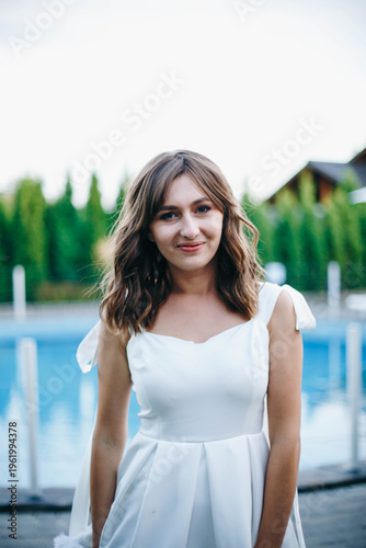 Young woman in white dress with feather hem posing near swimming pool, stylish summer portrait. Elegant feminine look, natural light, modern outdoor lifestyle scene with water reflections.