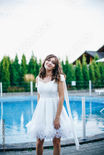 Young woman in white dress with feather hem posing near swimming pool, stylish summer portrait. Elegant feminine look, natural light, modern outdoor lifestyle scene with water reflections.