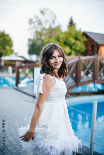 Young woman in white dress with feather hem posing near swimming pool, stylish summer portrait. Elegant feminine look, natural light, modern outdoor lifestyle scene with water reflections.