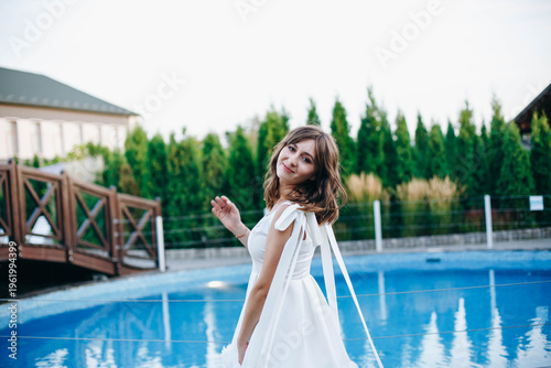 Young woman in white dress with feather hem posing near swimming pool, stylish summer portrait. Elegant feminine look, natural light, modern outdoor lifestyle scene with water reflections.