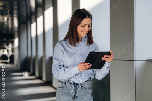 Wallpaper Mural Young woman with long brown hair wearing a light blue shirt holds a tablet while standing in a modern corridor with natural light streaming through windows Torontodigital.ca