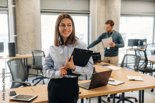 Wallpaper Mural Young woman in blue blouse holding a clipboard stands in modern office space with large windows, while a man in background reviews documents at a desk Torontodigital.ca