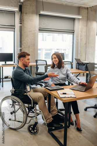 Wallpaper Mural Young man in wheelchair and woman in office attire collaborate at a desk with laptop and documents in a modern workspace with large windows and natural light Torontodigital.ca