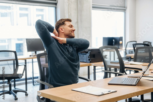Φωτογραφία Male office worker in blue sweater stretches in modern workspace with wooden des