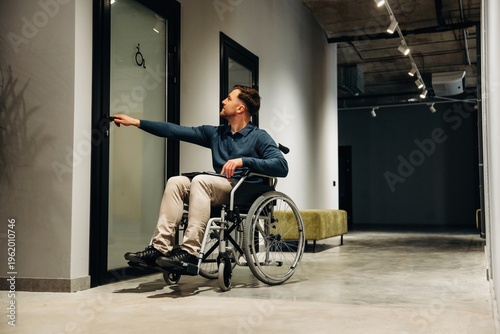 Young man in wheelchair reaching for door handle in modern interior space with concrete flooring and minimalist design elements visible in the background