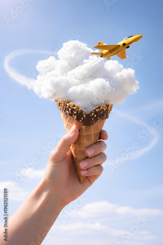 Surreal travel concept – yellow jet flying through cloud inside ice cream cone held by female hand against blue sky