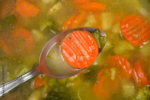A metal spoon holds a sliced cooked carrot over a pot of hot chicken and vegetable soup with broccoli and cauliflower