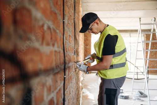 Wallpaper Mural Male construction worker in reflective safety vest using tools to install electrical wiring on exposed brick wall in a partially renovated building interior Torontodigital.ca