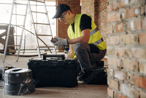 Wallpaper Mural Male construction worker in safety vest examines tools from a black toolbox while sitting on the floor of a building under renovation with a ladder nearby Torontodigital.ca