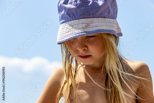 fair-haired girl with long hair in a blue panama against  blue sky, grimacing from the sun, sad, on the beach, medium plan, European, rest, summer day, relaxation, tourist, travel, summer, 6-8 years o