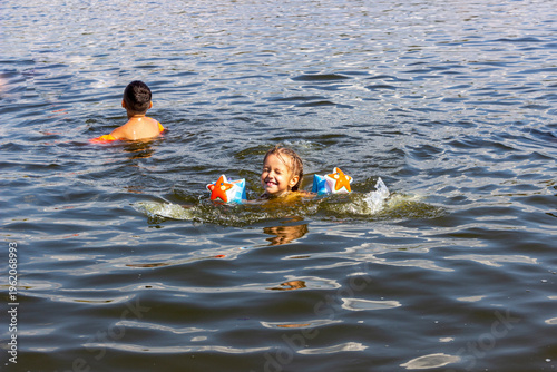 fair-haired girl in swimming armbands swims in river happily squints, laughs, splashes, portrait, muddies water, swims, childhood, rest, teenager, view straight, wet hair, pigtails, summer, day, sunny