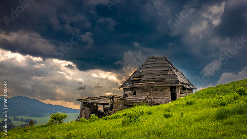 Abandoned wooden hut on green hillside under dramatic storm clouds, rural mountain landscape with moody sky and natural countryside scenery