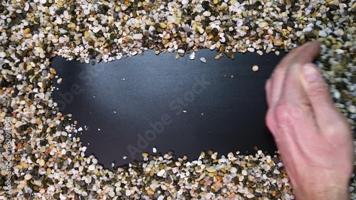 Man's hand moves aside small natural aquarium stones, leaving a strip of black background for text. Decorative stones for decor and aquariums.
