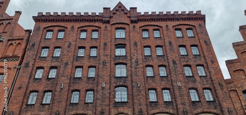 red brick facade from 1870 features multiple rows of arched windows and a crenelated roofline under a cloudy sky displaying classic industrial or Hanseatic architectural style and history