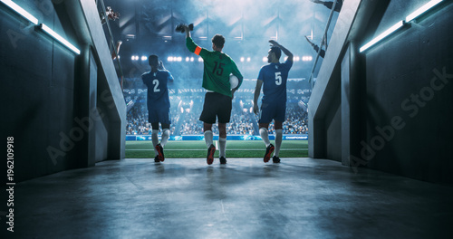 Three Male Soccer Professionals Stride From the Stadium Tunnel Onto the Illuminated Pitch, Led by a Goalkeeper Carrying the Ball, Acknowledging Spectators Before a High-profile Championship Match.