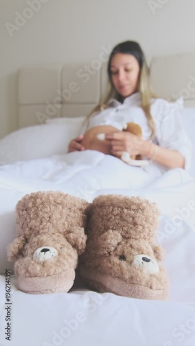 Expectant mother moisturizing her belly with lotion, with cute teddy bear baby shoes in the foreground