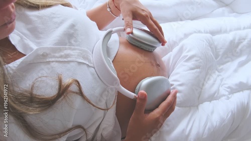 Expectant mother lying in bed holding headphones on her pregnant belly for the unborn baby to listen