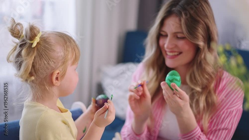 Mother and child paint Easter eggs together. Smiles fill the warm, bright room. Child holds a purple egg, mother a green one. They sit at a table with colorful tools