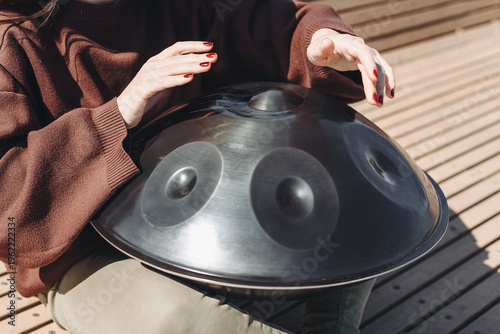 young caucasian woman playing black handpan with hands, ethnic metal drum instrument, sits on wooden bench of enbankment, beautiful relaxing music for meditation, close-up view of hands