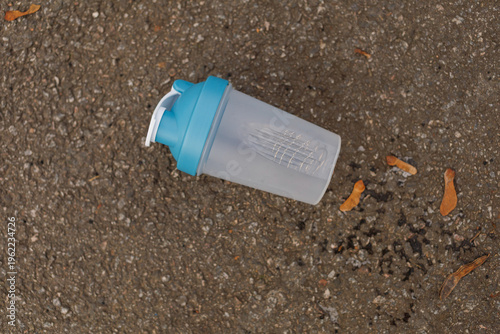A shaker bottle rests on the ground next to dry leaves in a city environment