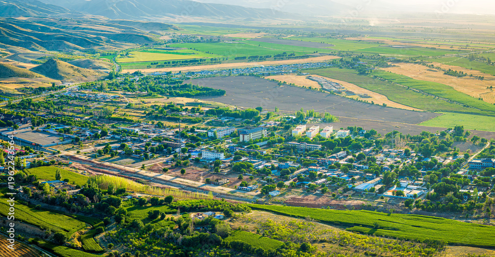 Fototapeta premium Aerial view of a peaceful village nestled in a lush valley with vibrant agricultural fields and rolling mountains in Xinjiang, China.