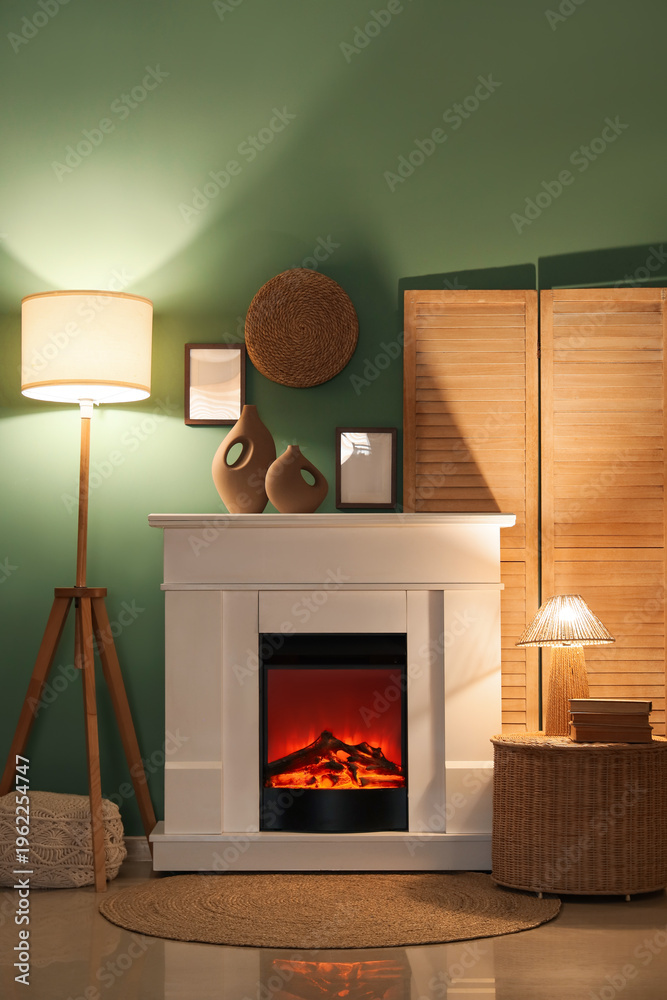 Fototapeta premium Interior of evening living room with folding screen, glowing lamps, wicker table and home decor on fireplace near blue wall