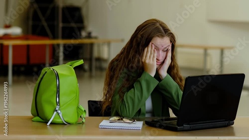 Stressed young woman in green blazer sits at desk with laptop and bright backpack, rubbing her face from fatigue and information overload, concept student burnout and online learning stress.