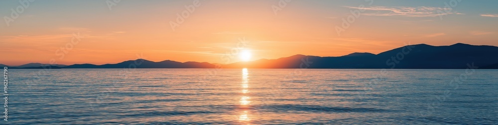 Fototapeta premium Sun setting behind distant mountains, reflecting orange light on the calm ocean surface. Serene nature scene at twilight