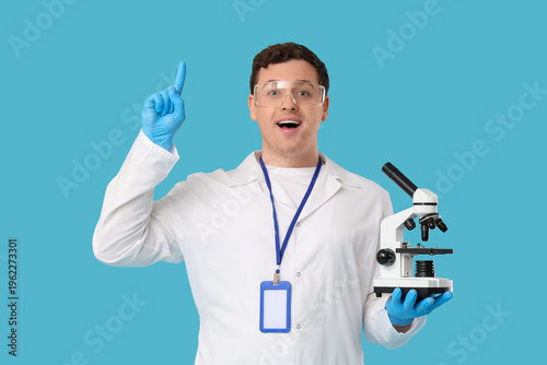Surprised young male scientist with microscope pointing up on blue background