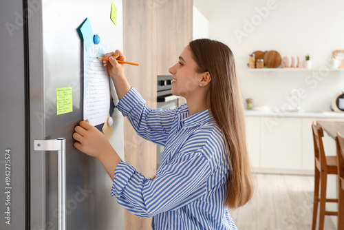 Young woman making to-do list on fridge in kitchen