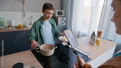 Over shoulder of two Caucasian teenagers using smartphone on stabilizer to film cooking video, boy stirring spaghetti in pot while girl recording in bright modern kitchen