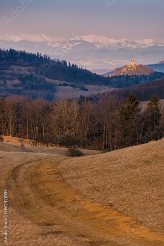 Scenic countryside landscape with a winding dirt road leading through hills towards a hilltop church. Snow-capped peaks of the Nizke Tatry in the background. Peaceful rural scene in Slovakia.