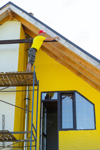 Cuadro en lienzo Worker balances on upper scaffold attaching horizontal plank under gable, yellow contemporary facade, critical construction moment, personal safety gear