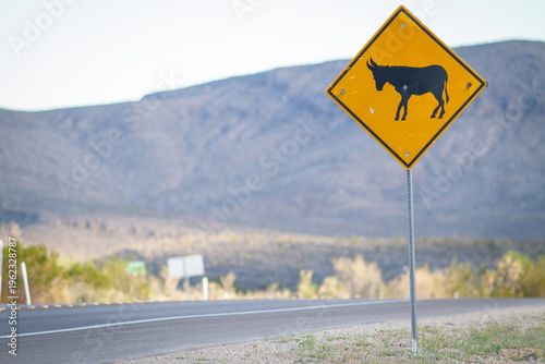 A donkey crossing road sign along an American southwest desert highway. A jackass against a yellow diamond signaling caution that large animal may be crossing the street at this location