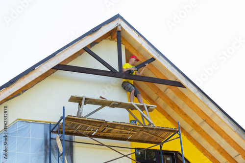 Картината върху платно Carpenter wearing helmet paints decorative crossbeam of gable from scaffolding, visible tools and equipment, yellow modern house, focus on safety