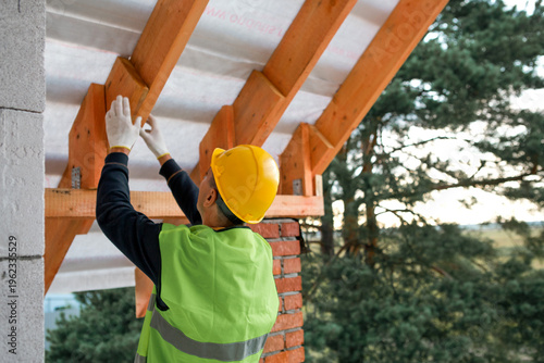 Photos Builder in helmet attaches roof rafter to bearing wall during house construction, timber roof frame assembly, active construction phase