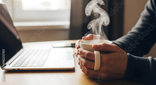 Person holding steaming hot coffee cup beside laptop on wooden desk in office