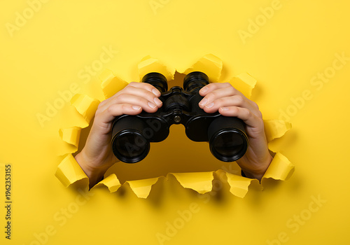 Hands holding binoculars through torn yellow paper background