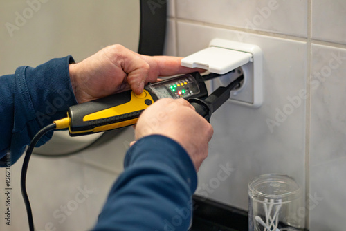 Electrician using a voltage tester to check electrical outlet in a modern bathroom with white tiles and a round mirror reflecting the scene